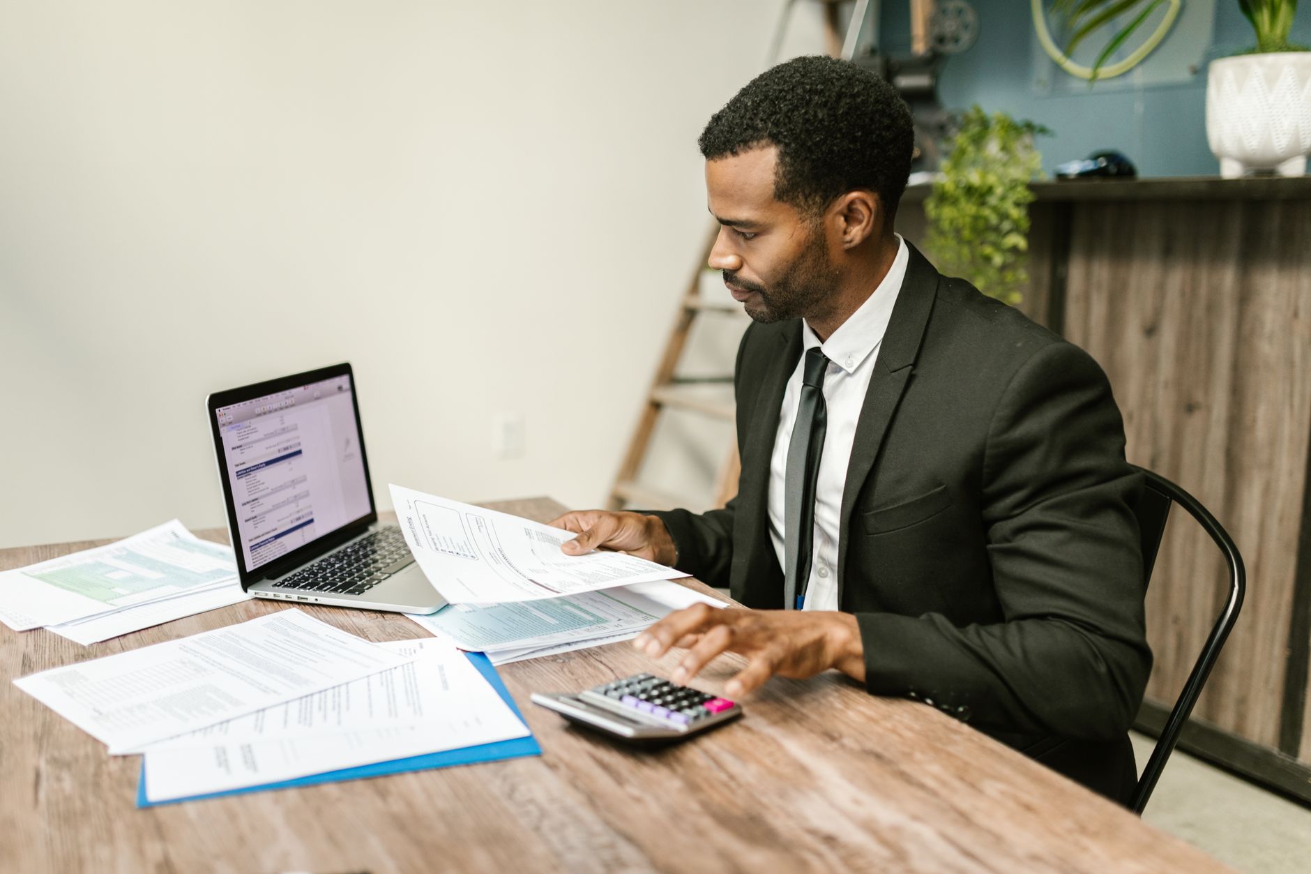 Homme en costume examinant un document et utilisant une calculatrice devant un ordinateur portable et des feuilles sur une table en bois.