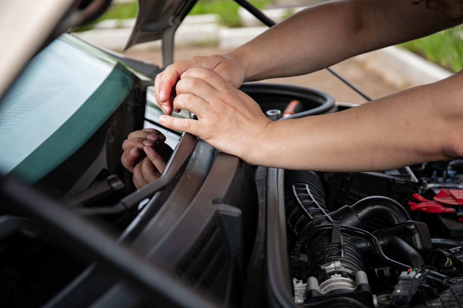 Mains travaillant sous le capot ouvert d’une voiture, près du pare-brise et du compartiment moteur.
