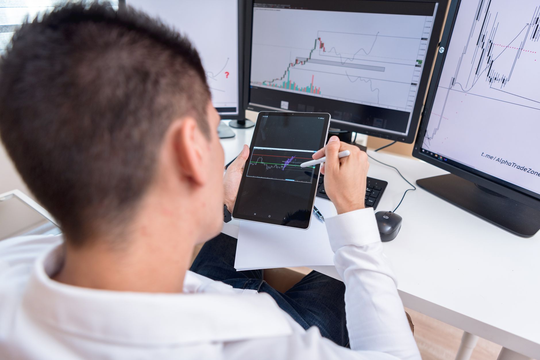 Un homme analyse des graphiques boursiers sur des écrans et une tablette à son bureau.