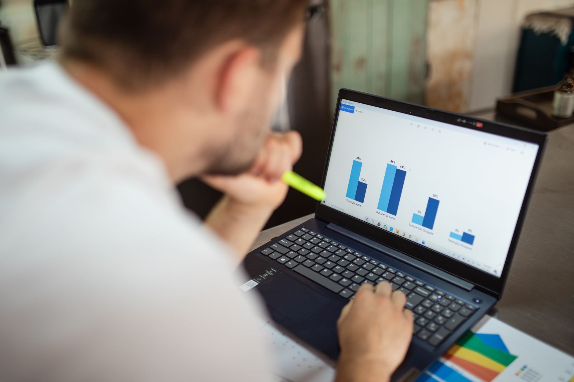 Un homme examine un graphique en barres sur un ordinateur portable, un surligneur à la main, devant un bureau avec un document imprimé.