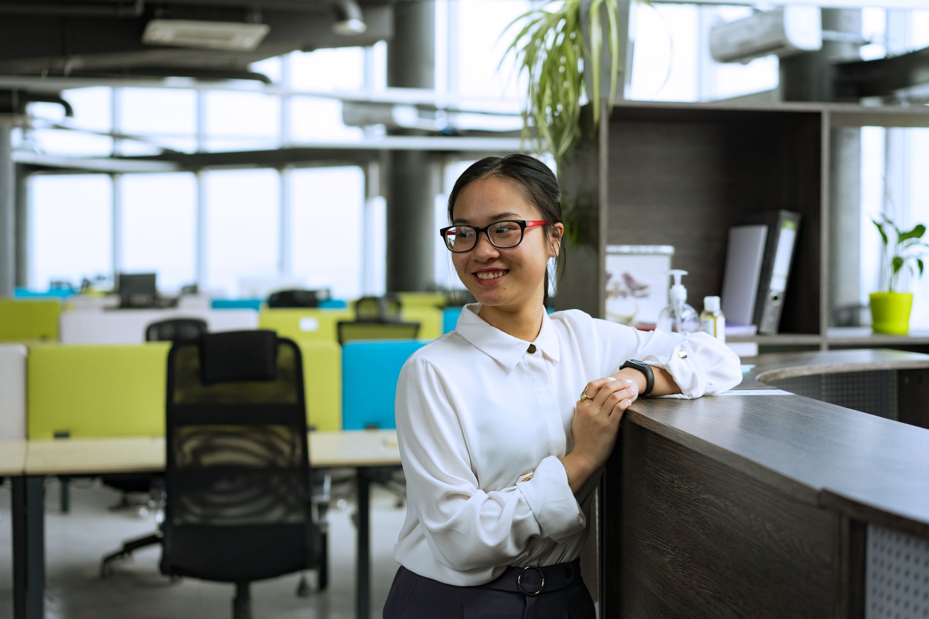 Une femme souriante en chemise blanche et lunettes, accoudée à un comptoir d’accueil dans un open space lumineux.