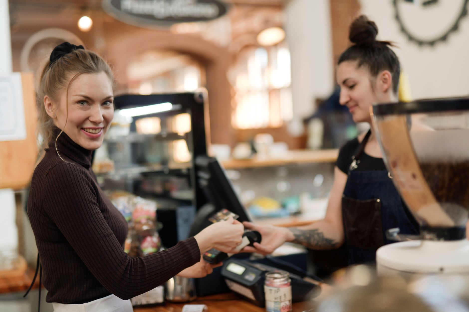 Une jeune femme souriante tend sa carte à un terminal de paiement au comptoir d’un café, face à une employée en tablier, près d’un moulin à café.