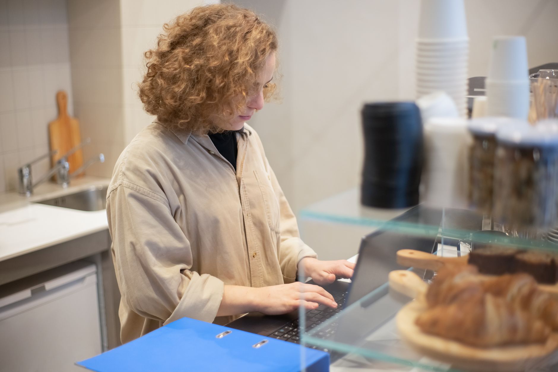 Une personne travaille sur un ordinateur portable derrière le comptoir d’un petit commerce, avec un classeur bleu et des viennoiseries en vitrine.